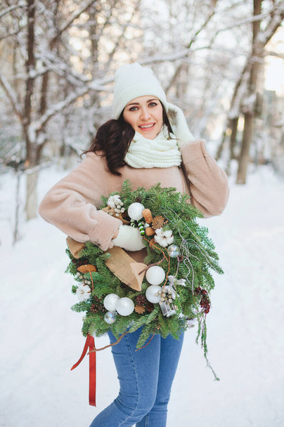 Νεαρή γυναίκα με εξωτερικά ενδύματα που φέρει διακοσμημένο στεφάνι Χριστουγέννων και στέκεται στο χιόνι την ηλιόλουστη χειμωνιάτικη μέρα - Φωτογραφία, εικόνα