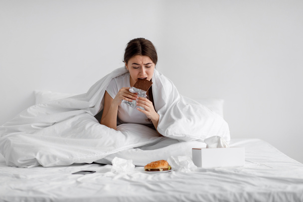 Грустный голодный кавказский юная леди ест шоколадный батончик на кровати с фаст-фудом дома, страдает от стресса - Фото, изображение