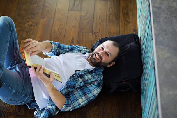 Portrét radostného mladého muže, jak čte knihu, zatímco sedí na podlaze ve svém obývacím pokoji.Student drží a čte knihu. - Fotografie, Obrázek