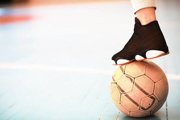 Un pie humano descansa sobre el balón en el suelo de hormigón. Foto de una pelota de fútbol y zapatillas en un piso de madera - Foto, Imagen