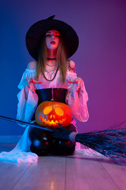 a girl in a witch costume for Halloween with a broom and a pumpkin in neon light - Valokuva, kuva