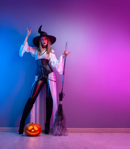 uma menina em um traje de bruxa para o Halloween com uma vassoura e uma abóbora em luz de néon - Foto, Imagem