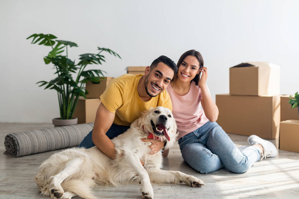 Full length portrait of happy young diverse couple with their dog posing on floor of new home on moving day - Photo, Image