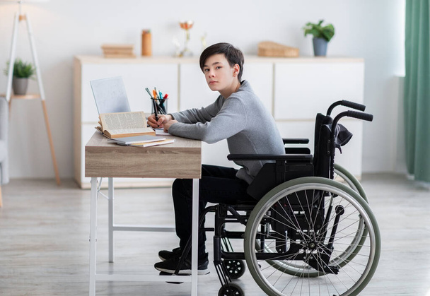 Tekerlekli sandalyedeki engelli genç evde bilgisayar ve kitaplarla online öğreniyor. - Fotoğraf, Görsel