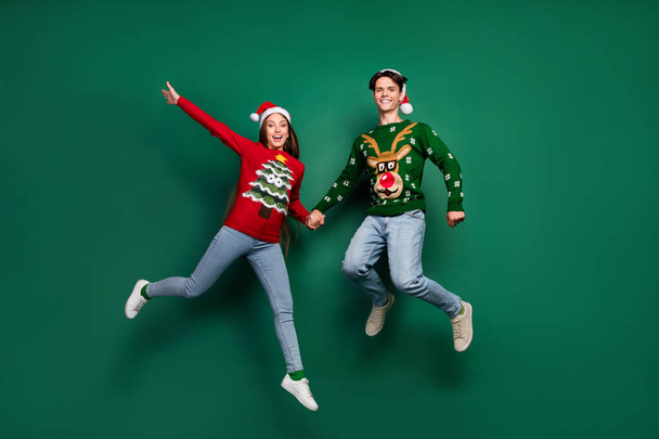 Photo of cheerful x-mas couple jump party wear santa hat ugly sweater isolated green color background - Φωτογραφία, εικόνα
