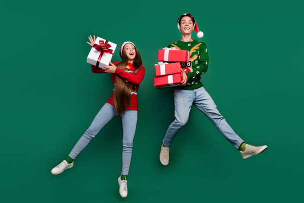 Photo of inspired two friends jump hold stack giftboxes wear santa hat ugly pullover isolated green color background - Foto, immagini