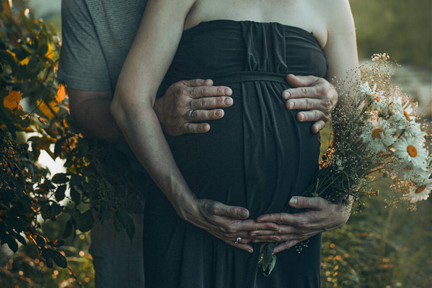 Pregnant woman and her husband hugging her tummy standing outdoors surrounded by nature. Pregnancy, expectation, motherhood concept - Фото, изображение