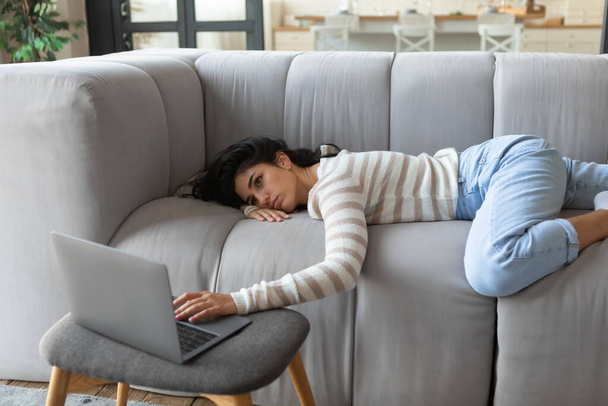 Уставшая молодая кавказка лежит на диване, пользуется ноутбуком, слишком измучена или скучает для работы в Интернете или учебы дома - Фото, изображение