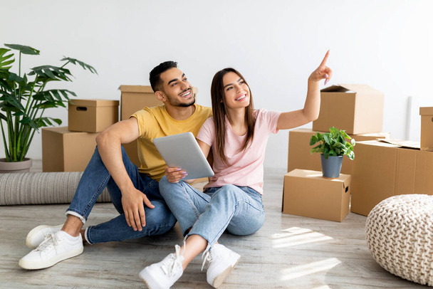 Happy interracial couple with tablet pc sitting on floor among carton boxes and planning design of their new apartment - Photo, Image
