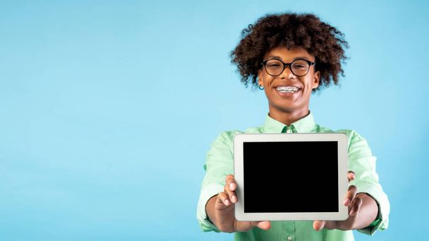 Веселый черный подросток показывает новый цифровой планшет с чистым экраном на голубом фоне, панорама, свободное пространство - Фото, изображение