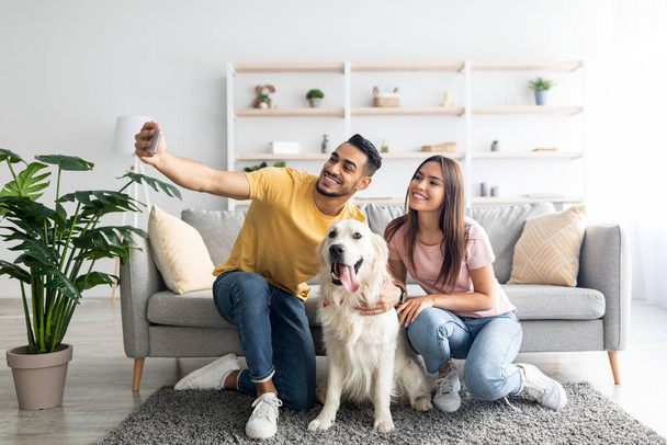 Happy interracial couple taking selfie with their dog while sitting on soft carpet at home, full length - Foto, Imagen