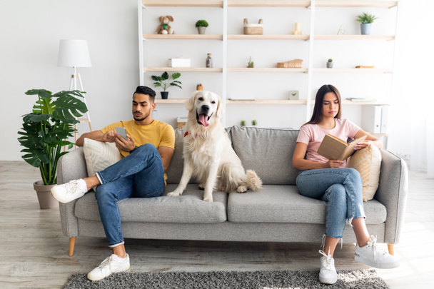 Joven pareja inrerracial que tiene conflicto, sentado en lados opuestos del sofá en casa, perro mascota entre ellos - Foto, imagen