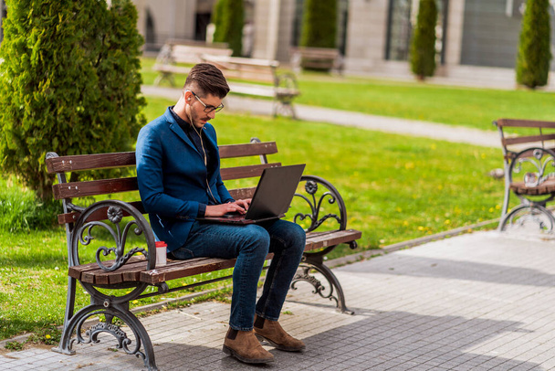 Schöner junger Manager beim Mittagessen, auf Holzbank sitzend und am Laptop arbeitend - Foto, Bild