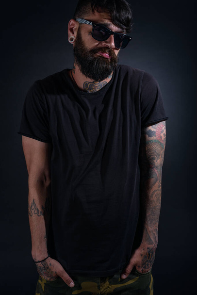Bearded male model with tattoos and hands in his pocket, dressed in a black t-shirt and sunglasses poses over black background. - Foto, Imagem