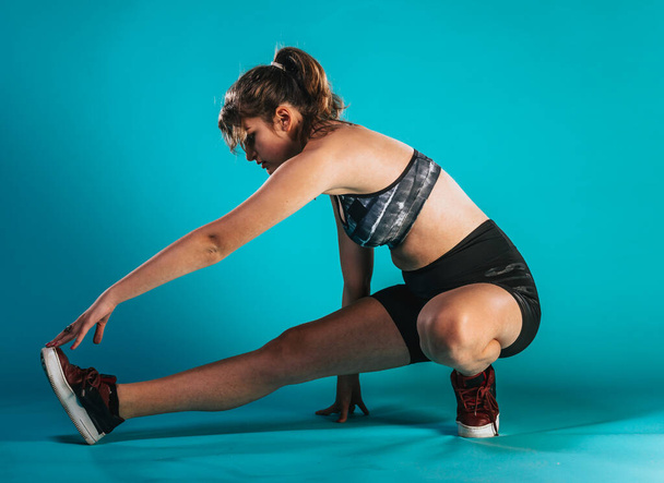 Eine junge Trainerin in einem sportlichen kurzen Top Stretch Beine auf blauem Hintergrund im Studio - Foto, Bild