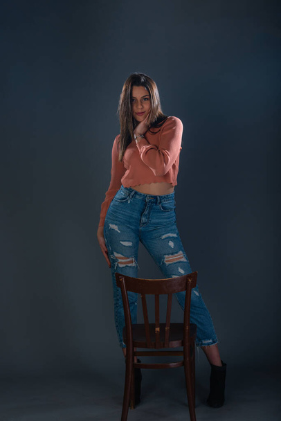 Female model with an orange crop top blouse and jeans posing in front of a wooden chair - Photo, image
