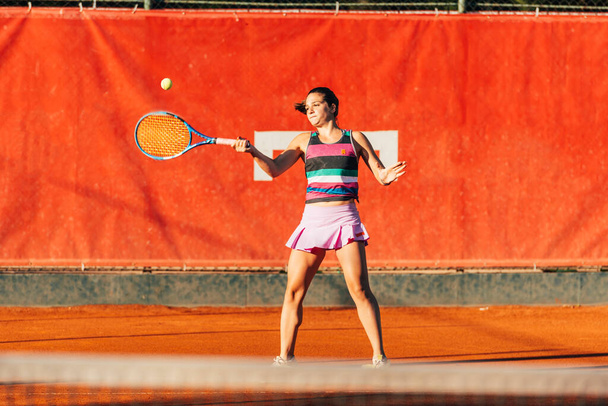 Μια νεαρή γυμνασμένη γυναίκα παίζει τένις σε ένα πορτοκαλί γήπεδο τένις νωρίς το πρωί - Φωτογραφία, εικόνα