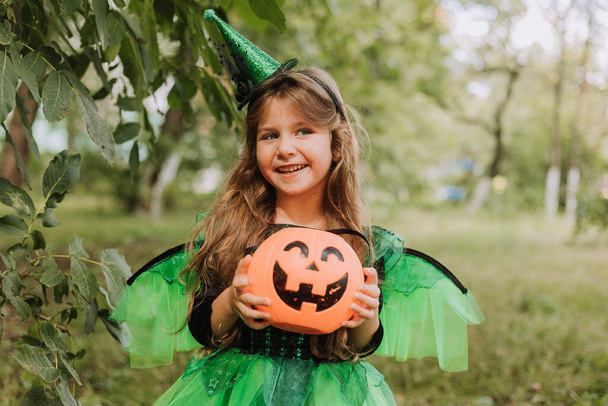 Милая маленькая девочка в костюме ведьмы на Хэллоуин держит корзину тыквы в парке. Ребенок на улице. Кошелек или жизнь. Концепция Хэллоуина - Фото, изображение