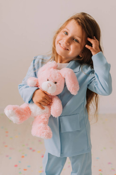 Mavi takım elbiseli, elinde pembe oyuncak tavşanla şirin bir kızın stüdyo portresi. Gri stüdyo geçmişinde poz veren güzel kız.  - Fotoğraf, Görsel