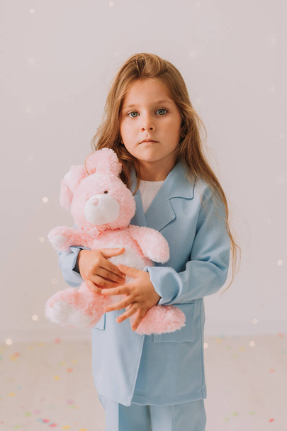 Studioporträt des niedlichen Kindermädchens im blauen Freizeitanzug mit rosa Spielzeughasen. Schönheit Mädchen posiert auf grauem Studiohintergrund  - Foto, Bild