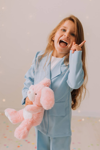 Studio portret słodkie dziecko dziewczyna w niebieskim garniturze casual gospodarstwa różowy króliczek zabawka. Piękna dziewczyna pozowanie na szarym tle studio  - Zdjęcie, obraz