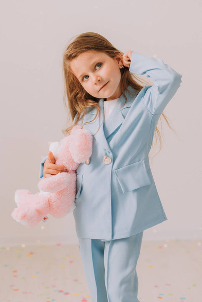 Studio portrait of cute child girl in blue casual suit holding pink toy bunny. Beauty girl posing on gray studio background  - Fotó, kép
