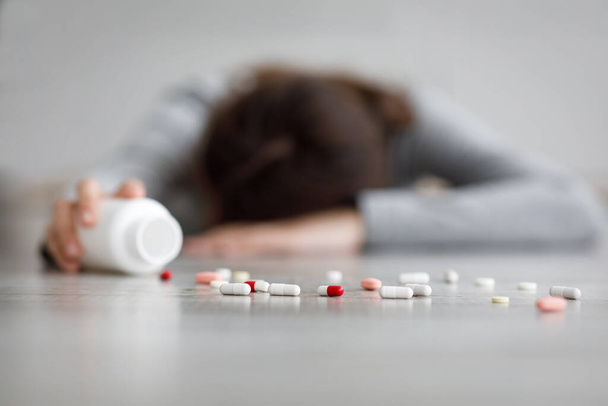 Unrecognizable young caucasian female lying on floor with jar and scattered pills on floor, committing suicide - Foto, Imagem