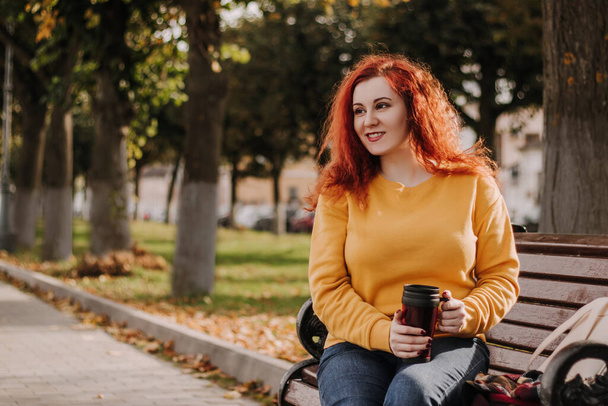 Portret van een jonge roodharige vrouw die in het park op de bank zit met een kop koffie. De dame draagt een gele sweater en houdt een herbruikbare beker vast. Lifestyle op een zonnige herfstdag. - Foto, afbeelding