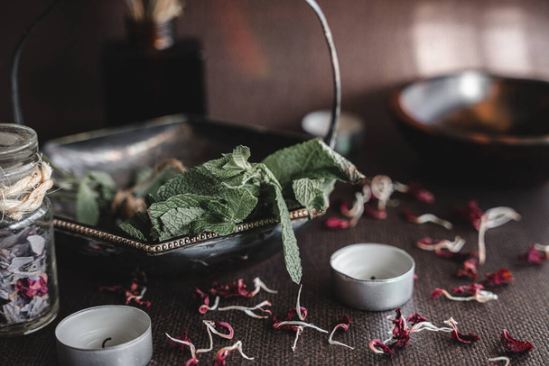 Bunch of fresh mint in a metal rustic vintage old bowl on a wiccan witch altar. Spell ingredients on a table with white candles and random scattered things in the background - Photo, Image