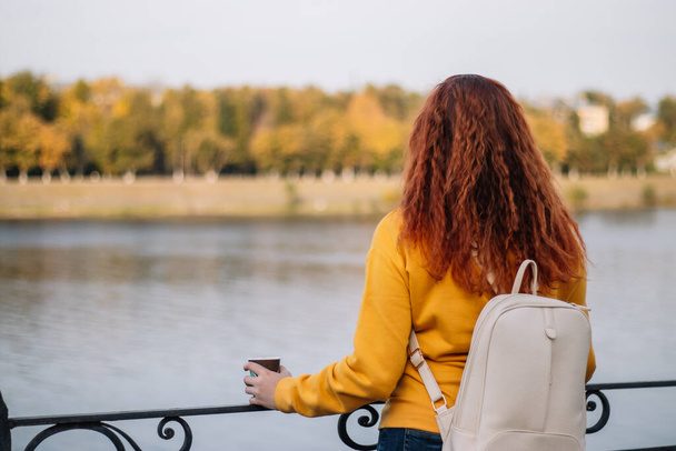 Back view of red-haired woman. Lady with white backpack enjoys autumn view and drinks coffee from paper cup. Eco-friendly lifestyle, copy space. - Photo, Image