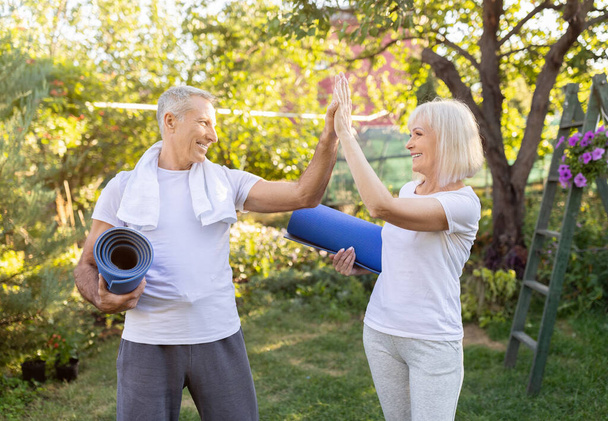 Active mature couple standing with yoga mats and giving high five each other after training in garden outdoors - Photo, Image