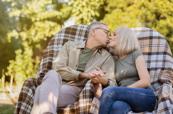 Starsza para małżeńska odpoczywa na świeżym powietrzu i całuje się, ciesząc się wakacjami siedząc na wiklinowych krzesłach w ogrodzie - Zdjęcie, obraz