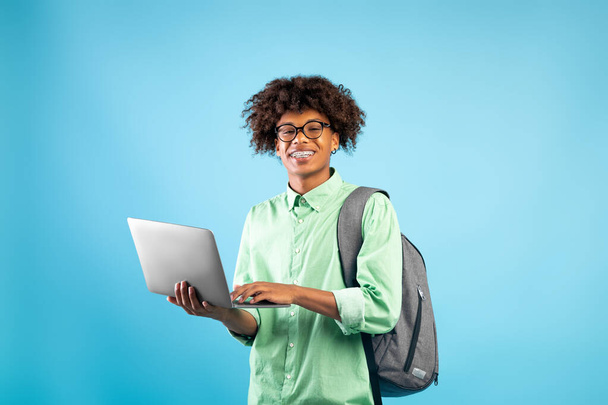 Online onderwijs concept. Happy zwart student guy met rugzak en laptop computer, staande over blauwe achtergrond - Foto, afbeelding