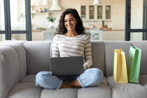 Verkkokaupoissa. Nuori nainen kannettavan tietokoneen ja ostoskassit tilaamalla tavaroita verkossa istuessaan sohvalla kotona - Valokuva, kuva