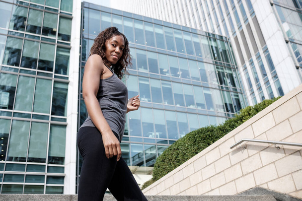 Фитнес-взрослая темнокожая женщина позирует в деловой среде. На ней чёрные леггинсы и серая футболка без рукавов.. - Фото, изображение