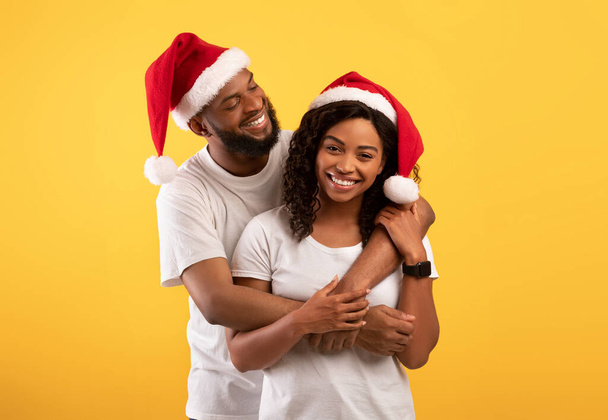 Loving Afrikaans Amerikaans paar in Santa hoeden knuffelen op gele studio achtergrond, vieren Nieuwjaar of Kerstmis - Foto, afbeelding