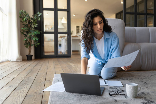 Очарованная молодая женщина-фрилансер, работающая в Интернете, просматривающая финансовые документы, использующая ноутбук в помещении, копирующее пространство - Фото, изображение