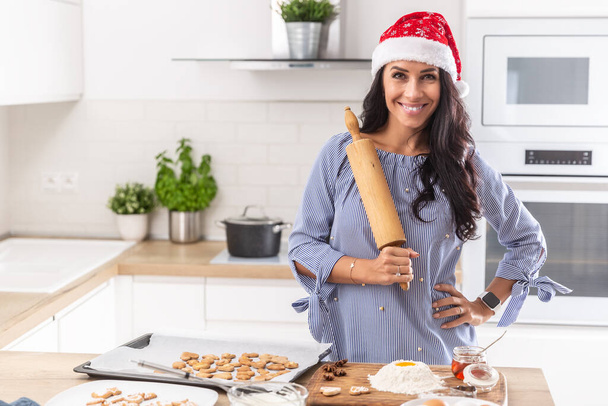 Christmas mother wears a christmas hat and holds the roller happily while baking chrismas cookies in her kitchen. - Foto, Imagen