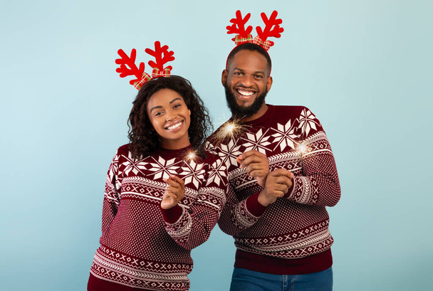 Время празднования. Радостная черная пара в оленьих рогах и свитерах, празднующих Рождество или Новый год с искорками - Фото, изображение