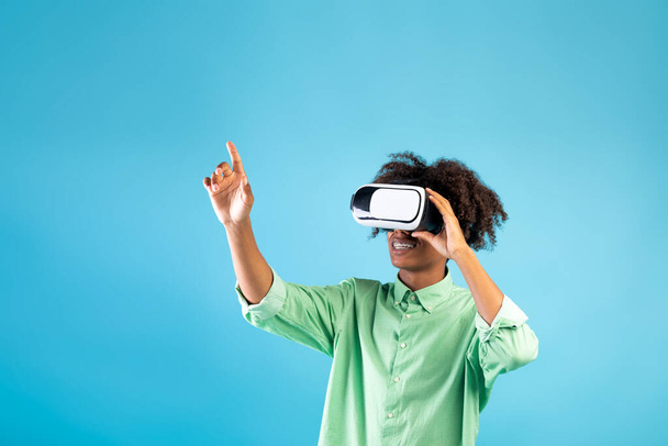 Monde virtuel en simulation 3D. noir adolescent guy dans vr lunettes jouer et essayer de toucher quelque chose sur bleu fond - Photo, image