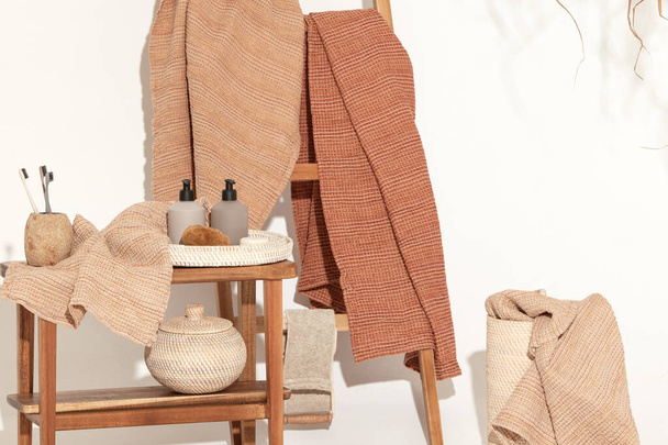 Natural waffle linen towels in earth tones on wood bench and towel ladder with bamboo toothbrushes, rattan baskets, and soap dispenser. Daily body care, spa and wellness zero waste bathroom concept  - Фото, изображение