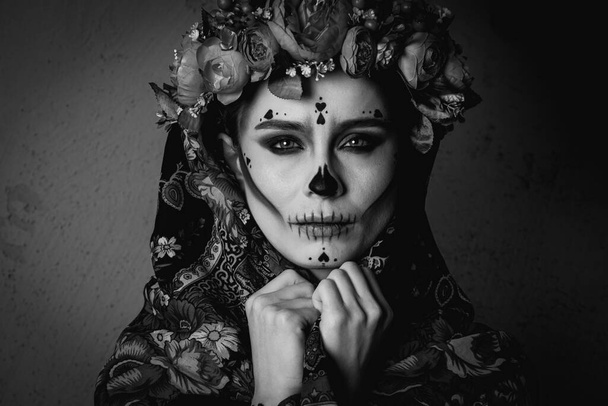 foto en blanco y negro de una joven con maquillaje para el Día de los Muertos con una corona de flores en la cabeza, con un encaje con flores bufanda se ve recta. El concepto de Halloween o La Calavera Katrina. - Foto, Imagen