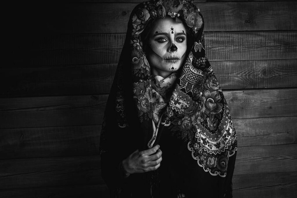 ασπρόμαυρη φωτογραφία ενός νεαρού κοριτσιού με μακιγιάζ για την Ημέρα των Νεκρών με ένα στεφάνι από λουλούδια στο κεφάλι του, φορώντας μια δαντέλα με φουλάρι λουλούδια φαίνεται ευθεία. Η έννοια του Halloween ή La Calavera Katrina. - Φωτογραφία, εικόνα