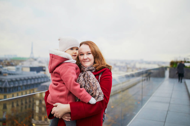 Young woman holding adorable todler girl while enjoying the view from Parisian rooftop terrace. Mothe and daughter in Paris, France - Fotografie, Obrázek