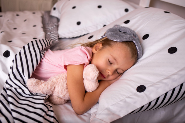 a beautiful little girl is sleeping in pajamas and a sleep mask with a soft toy on the bed in the bedroom - Photo, Image