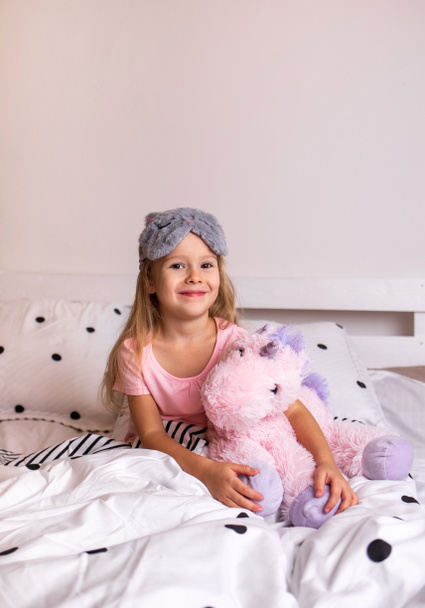 gelukkig klein blond meisje in pyjama zitten op het beddengoed op het bed in de kamer - Foto, afbeelding