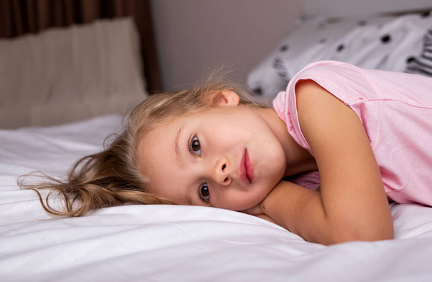 грустная маленькая блондинка лежит на белом постельном белье на кровати - Фото, изображение