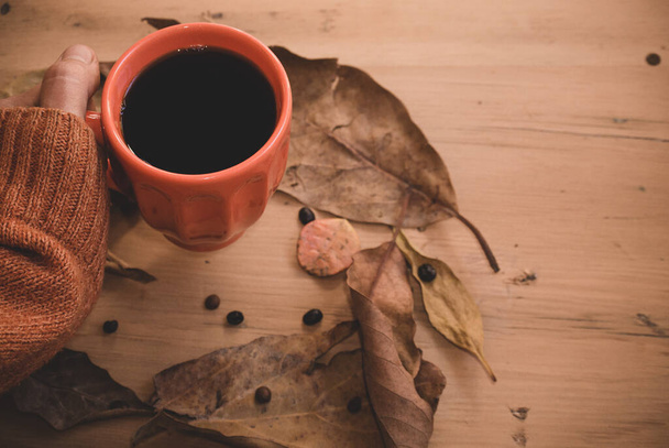 αρσενικό χέρι κρατώντας ζεστό καφέ, πορτοκαλί πουλόβερ και φύλλα του φθινοπώρου σε ένα παλιό ξύλινο φόντο. Αντίκες. Φθινοπωρινή έννοια χαλάρωσης. αντίγραφο χώρου - Φωτογραφία, εικόνα