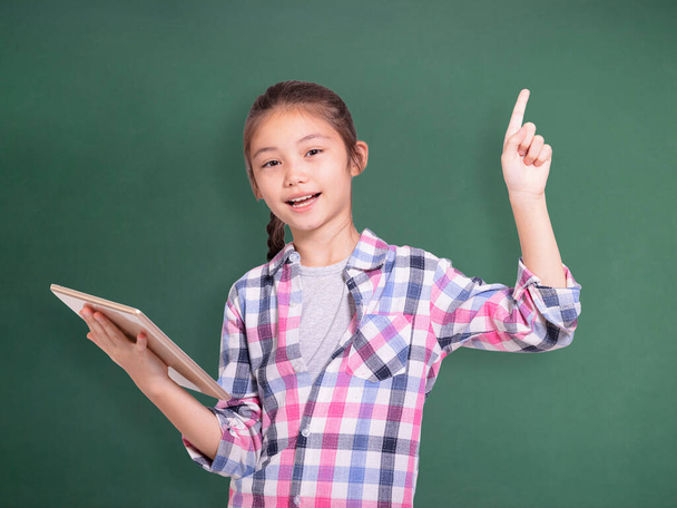Gelukkig student meisje holding tablet en wijzen upside.Isolated op groen schoolbord achtergrond. - Foto, afbeelding