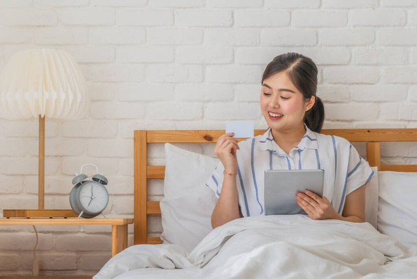 young asian woman lying on bed with tablet and blank credit card on hand making shopping and payment online looking at camera - 写真・画像
